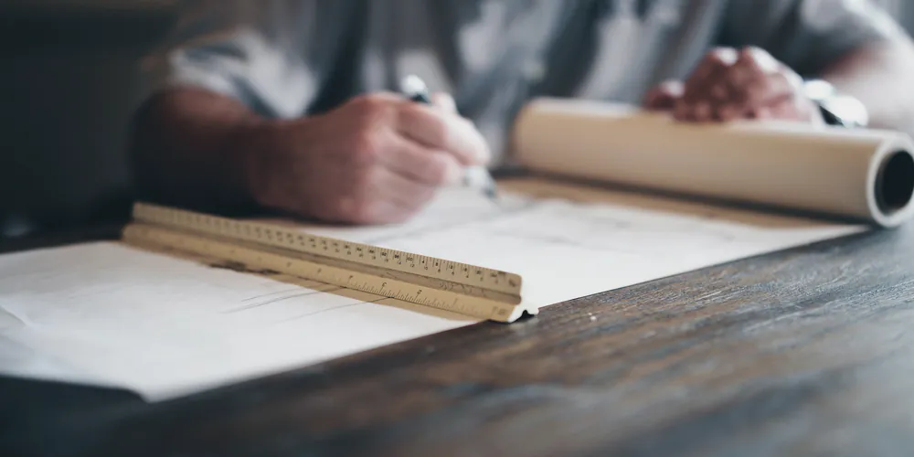 A person signing a document on a timber desk beside rolled plans