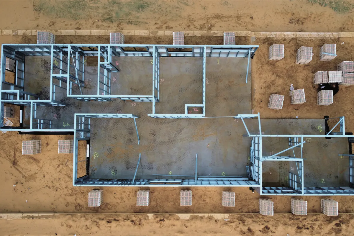 Steel wall frames standing on a new concrete slab, seen from above, with bricks stacked round it