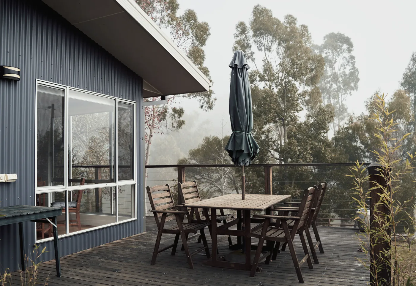 A timber deck with an outdoor table beside a dark clad house, gum trees in the mist beyond