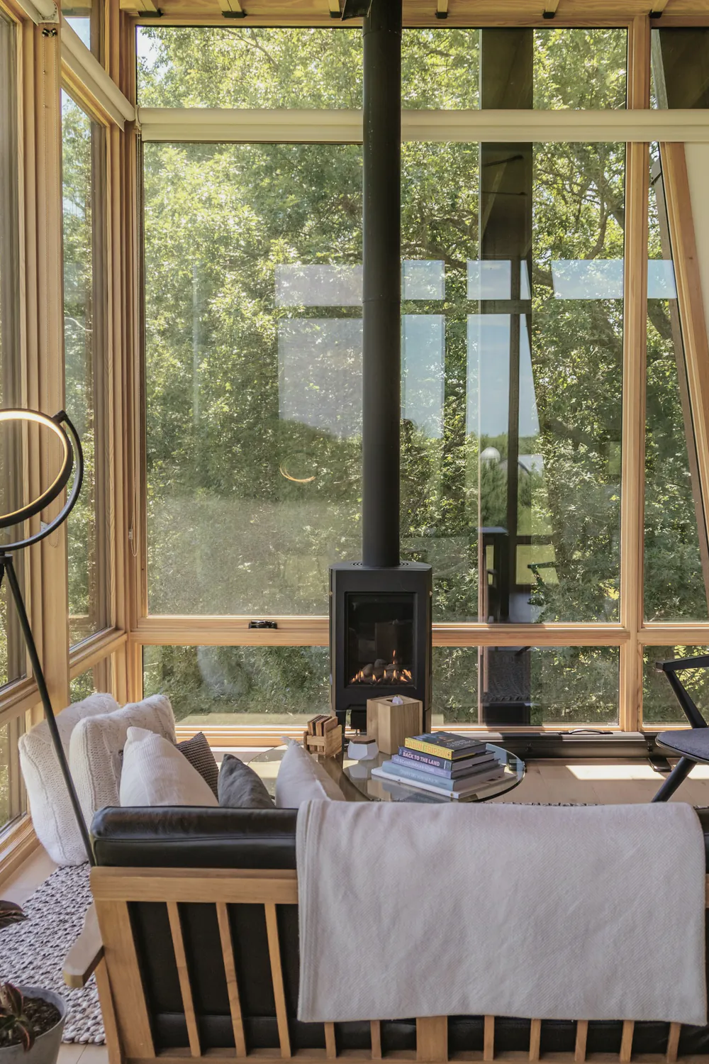A living room with a wood stove and full height glass on two sides looking into trees