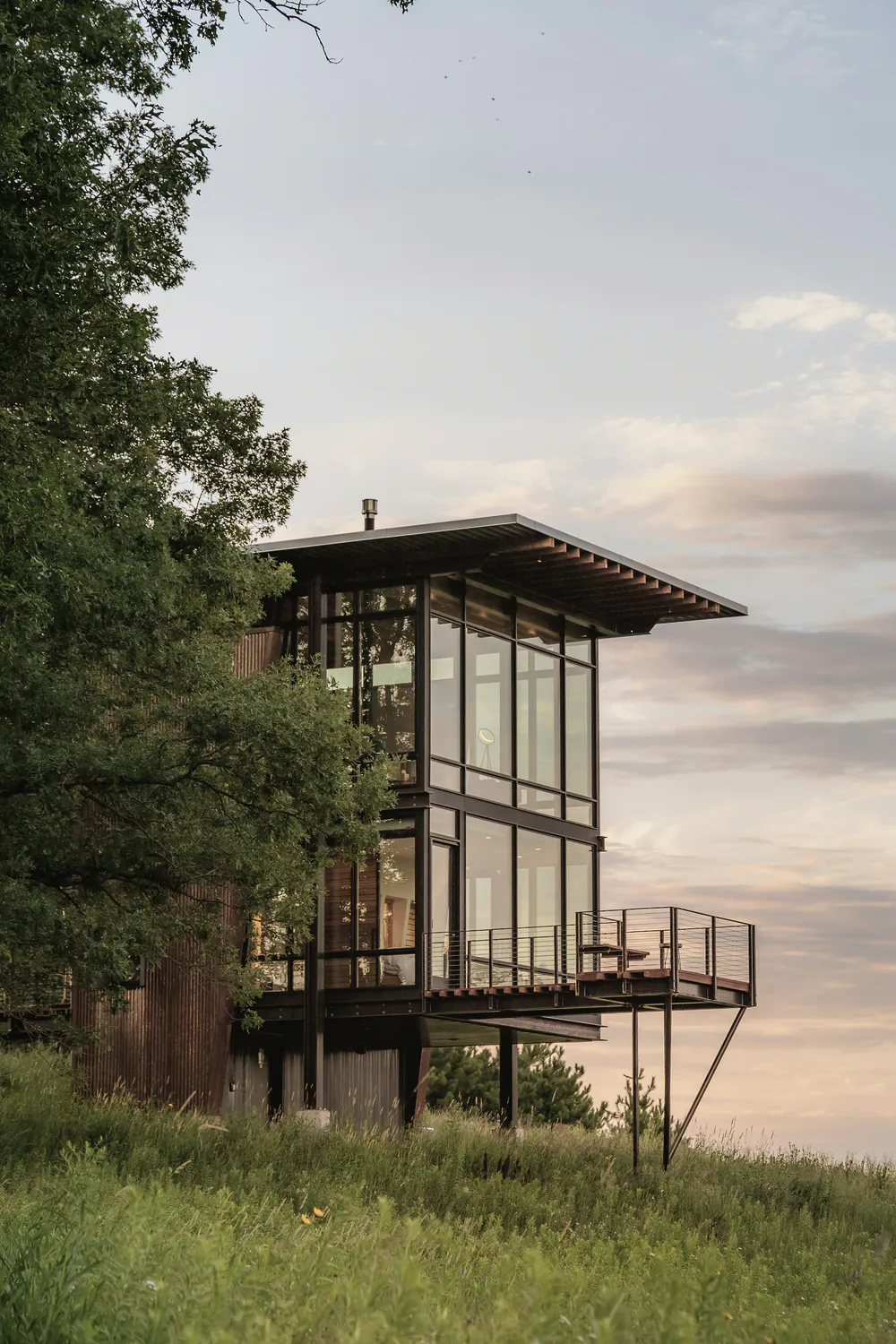 A glass and timber house lifted on steel piers among trees at dusk