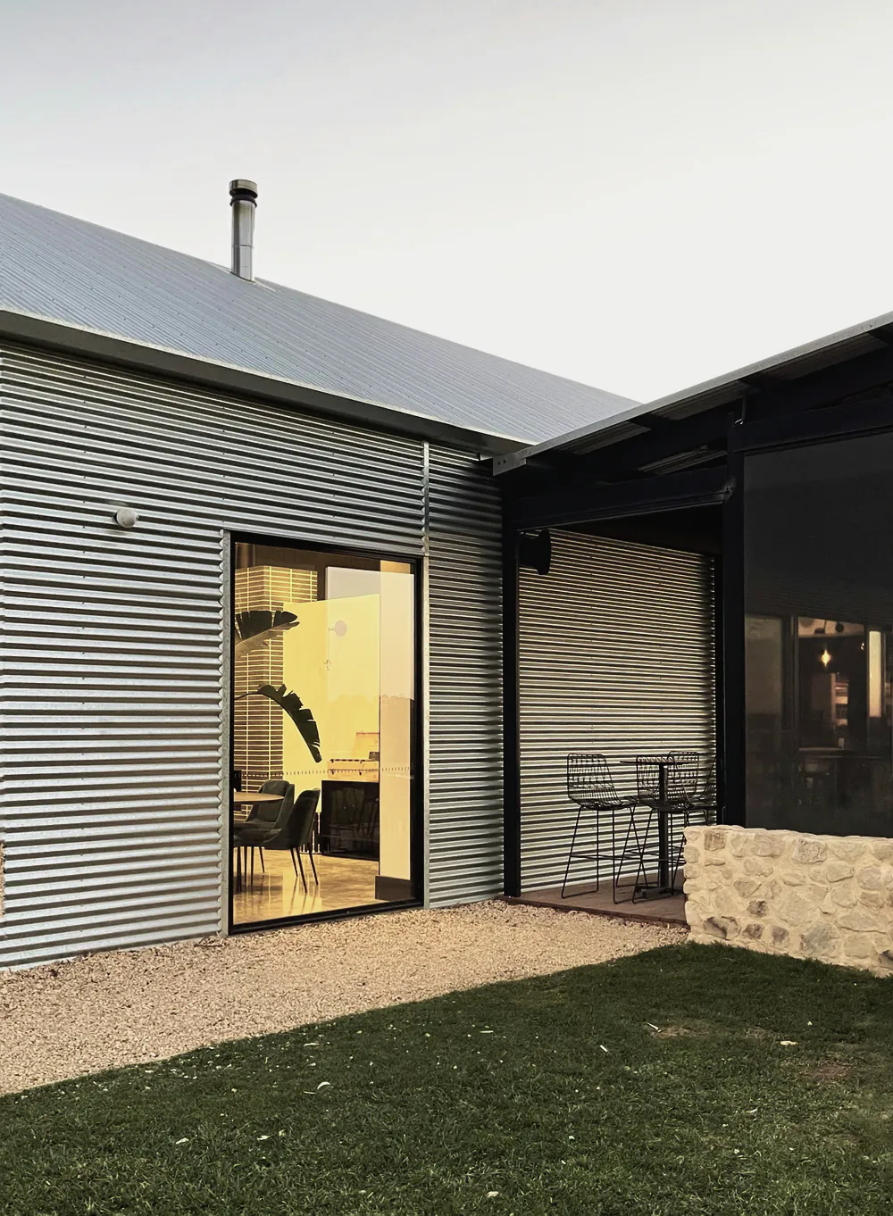 A corrugated iron house with a lit glass door opening onto gravel and lawn