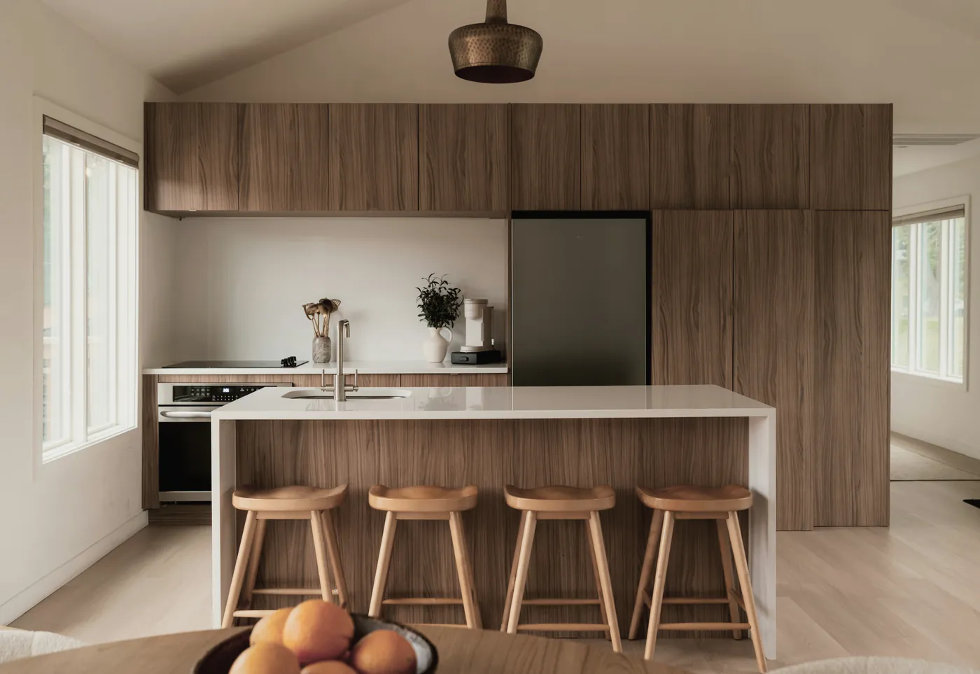 A walnut and white kitchen with four timber stools under a brass pendant, a bowl of oranges in front
