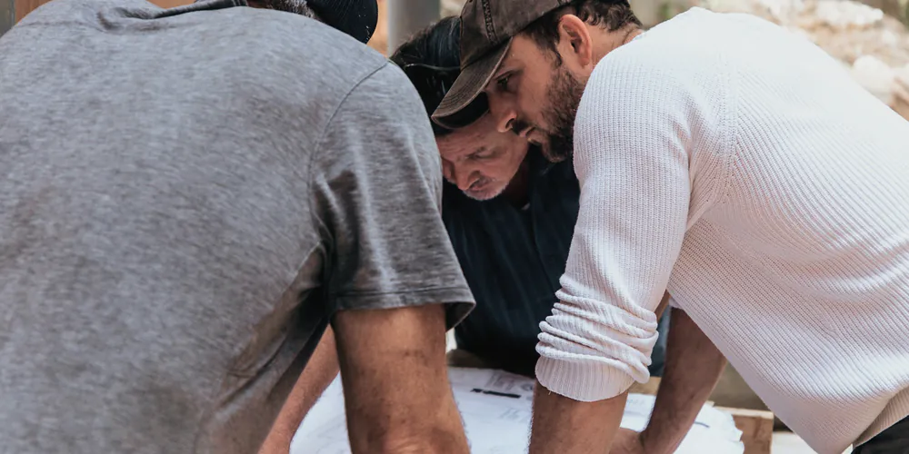Three builders leaning over a set of plans on a timber bench on site