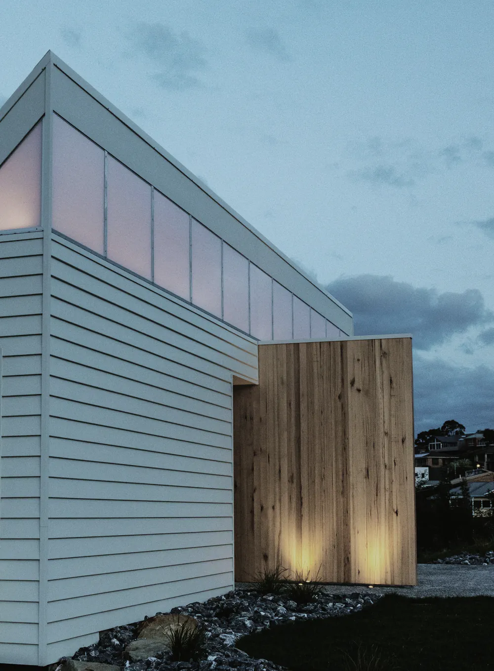 A white clad gable extension with a lit timber entry at dusk