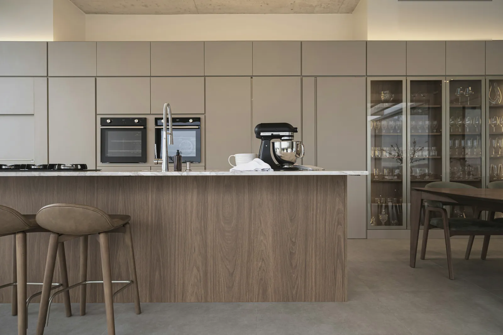 An oak kitchen island with a stone top and stools, glass fronted cabinets behind