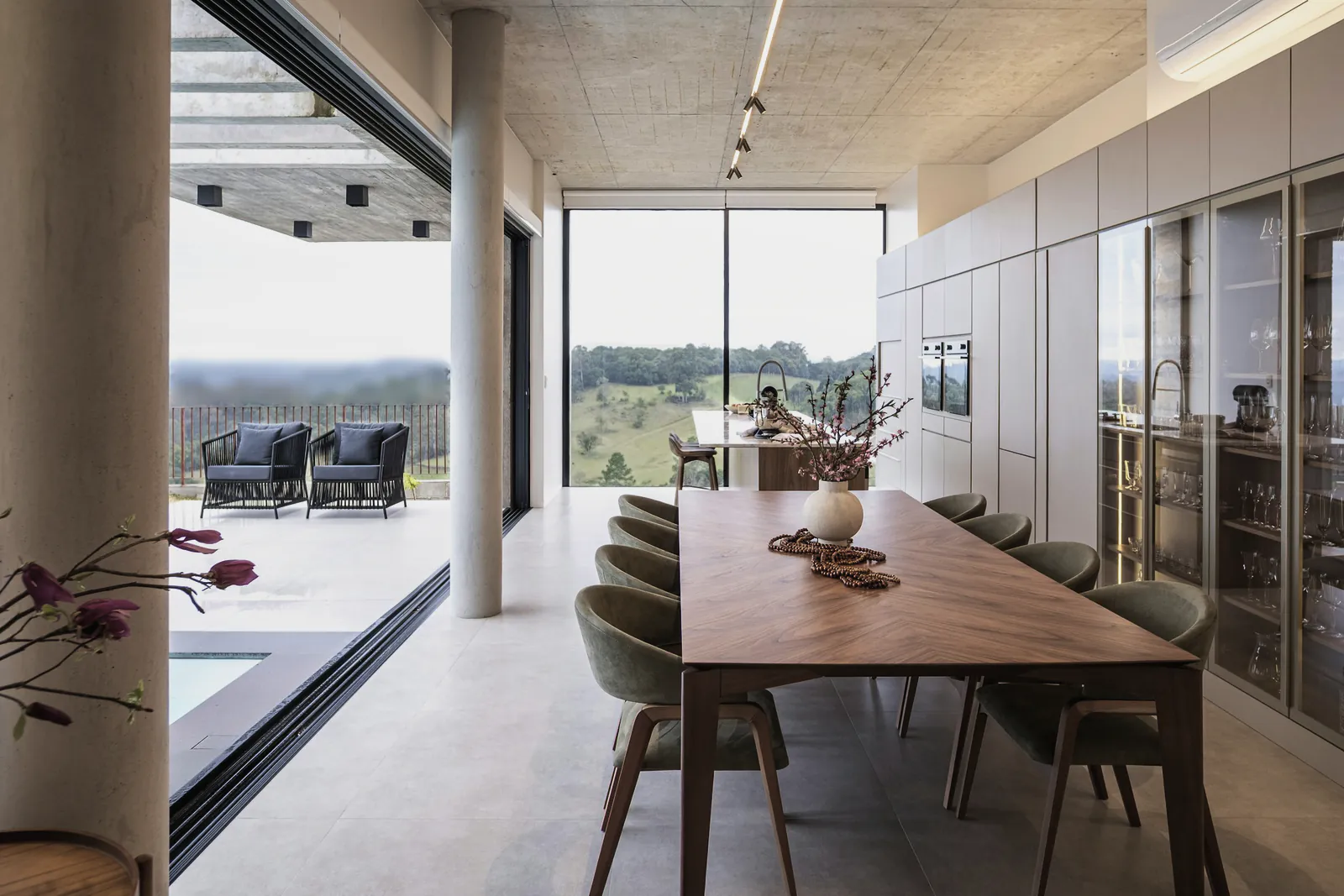 A long timber dining table under a concrete ceiling, the valley beyond open glass doors
