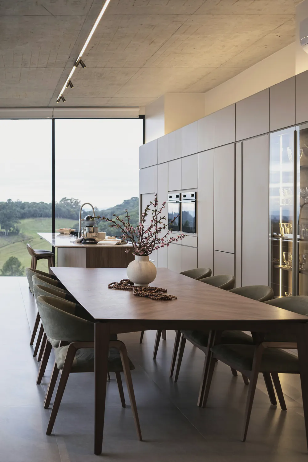 A long timber dining table under a board formed concrete ceiling, the valley beyond the glass