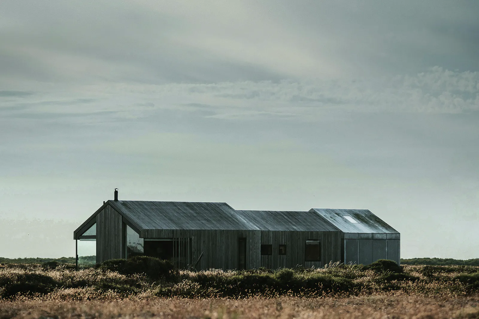 A long, low house clad in grey corrugated iron on open grassland under a pale sky