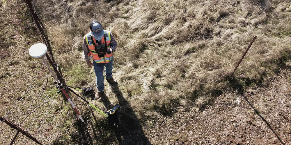 A surveyor with a GPS rover on a dry tussock block beside a wire fence, seen from above