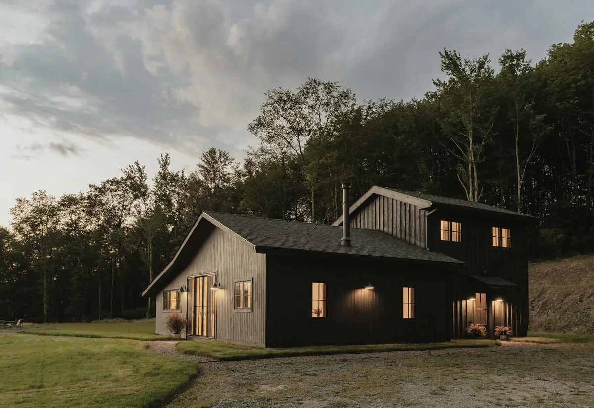 The two storey wing of a black timber house at dusk, its windows lit below a forest