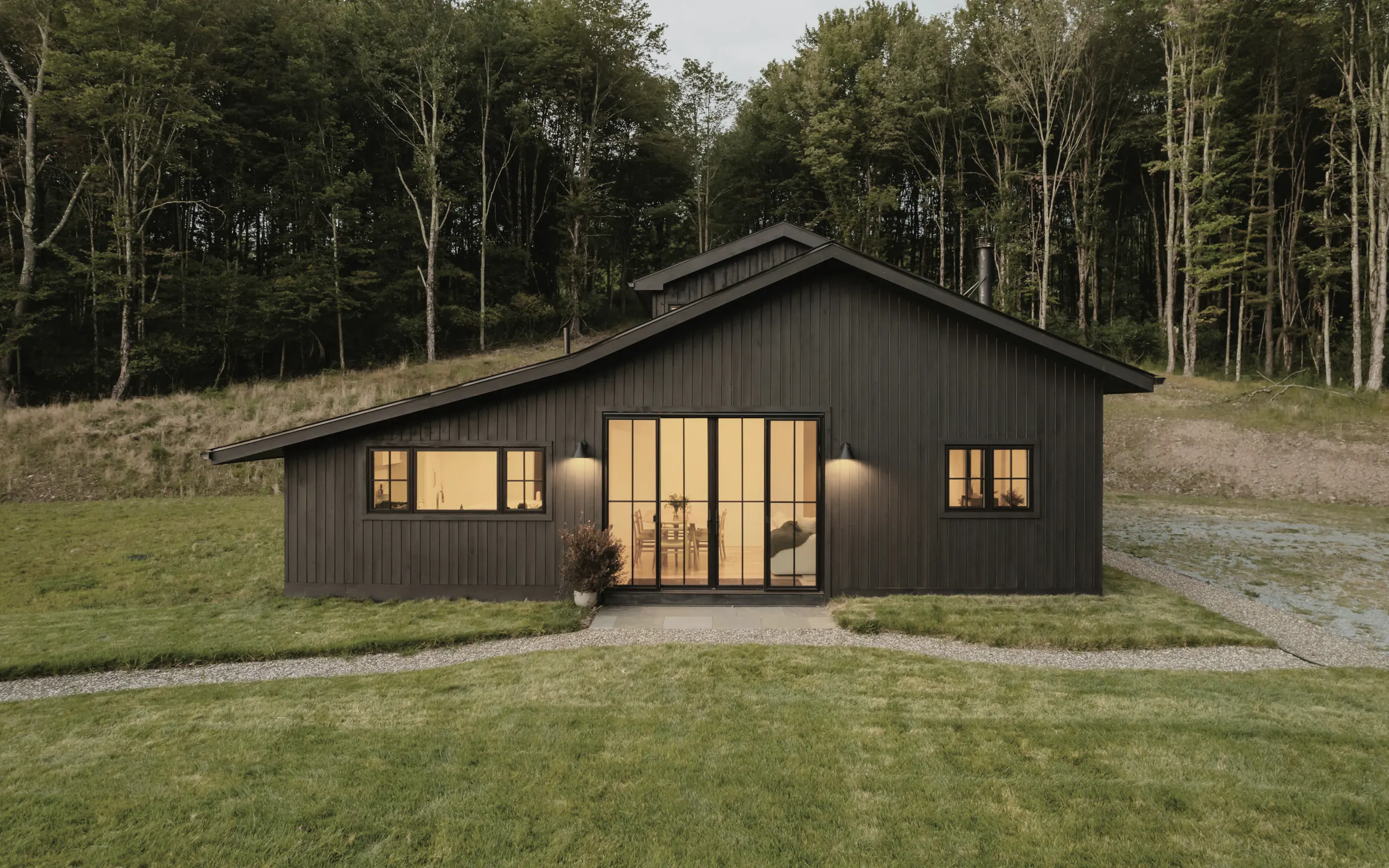 A black timber house with a gabled roof and lit glass doors on a mown lawn below a forest