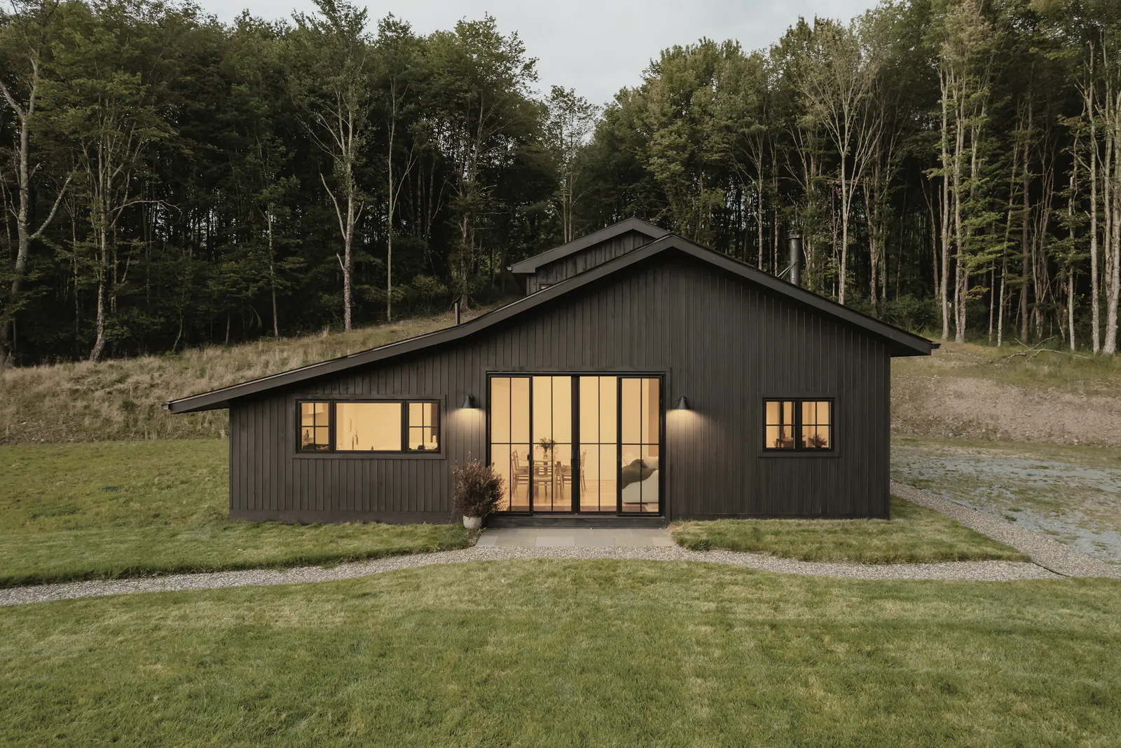 A black timber house with a gabled roof and lit glass doors on a mown lawn below a forest